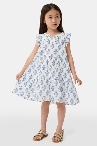 A young girl with long brown hair wears the Roller Rabbit Kids Rosa Ditsy Floral Pami Dress, made from cotton voile with blue-green block print and ruffled sleeves, standing against a plain light background, looking slightly to the side.