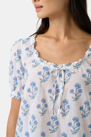 A woman with long dark hair wears the Roller Rabbit Rosa Floral Quincy Top, a cotton voile blouse with puffed sleeves, ruffled tie neckline, and matching blue flower patterns as part of a coordinating set.