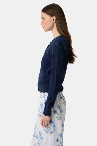 A woman wears the Roller Rabbit Pointelle Rosella Cardigan, a navy blue button-up with heart-shaped buttons, paired with white pants featuring a blue floral print, standing against a plain light background.