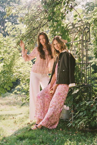 A woman stands barefoot against a plain white background wearing the Priya Floral Alvie Set by Roller Rabbit—a matching pink pajama set featuring a floral pattern, short-sleeve button-up top, and loose pants crafted from washable silk loungewear.