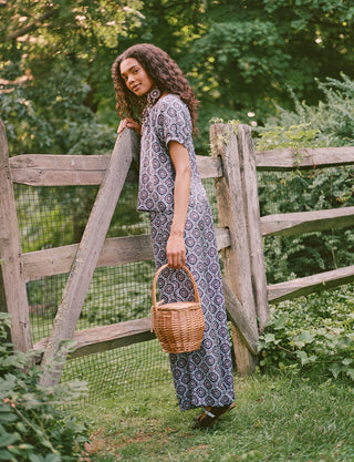 A woman stands by a wooden gate, holding a wicker basket in one hand. Outdoors amid greenery and trees, she gazes to the side with a smile, dressed in elegant Roller Rabbit Amelia Paige Pants.