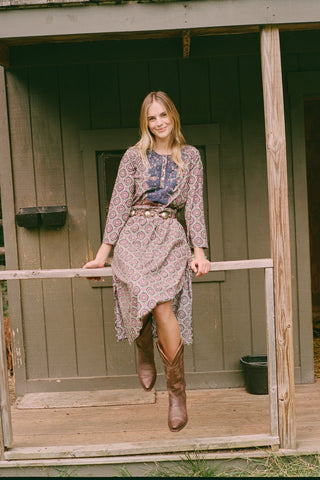 Wearing the Amelia Inez Long Kurta by Roller Rabbit, a woman with brown boots sits on the railing of a wooden porch, smiling, with a rustic building in the background.