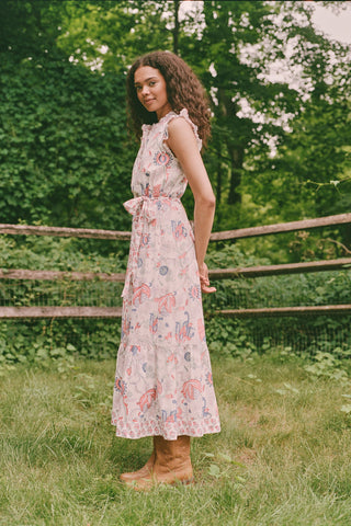 A person in the Amanda Nolan Dress by Roller Rabbit—a patterned, sleeveless, light-colored cotton maxi—stands on grass, holding out one side of the skirt. A wooden fence and lush greenery form the background; their face is not visible.