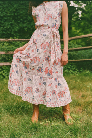 A person in the Amanda Nolan Dress by Roller Rabbit—a patterned, sleeveless, light-colored cotton maxi—stands on grass, holding out one side of the skirt. A wooden fence and lush greenery form the background; their face is not visible.
