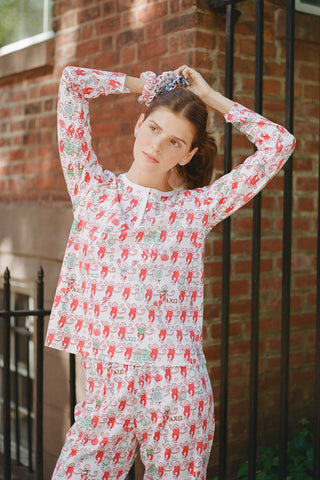A woman stands outside with her arms crossed, wearing the Roller Rabbit Alpha Chi Omega Monkey Pajamas—a white Henley top and elastic waist pajama set featuring playful red and pink monkey patterns. She holds a matching quilted bag and carries a small heart-shaped blue and white polka dot purse.