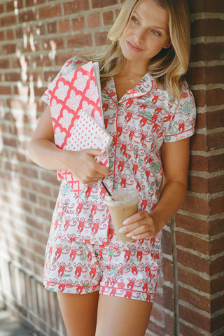 Wearing Roller Rabbit’s Alpha Chi Omega Monkey Polo Pajamas in a playful print, a woman leans against a brick wall, smiling softly as she holds two pink and white patterned notebooks and an iced coffee with a straw.