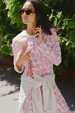 A woman wearing Roller Rabbit Gamma Phi Beta Monkey Pajamas—featuring a white and pink pattern with an elastic waistband—stands outdoors in front of greenery, holding a phone to her ear, a tumbler, and keys. A sweatshirt is tied around her waist, and she wears sunglasses.