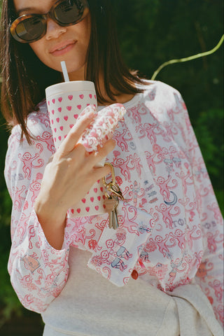 A woman wearing Roller Rabbit Gamma Phi Beta Monkey Pajamas—featuring a white and pink pattern with an elastic waistband—stands outdoors in front of greenery, holding a phone to her ear, a tumbler, and keys. A sweatshirt is tied around her waist, and she wears sunglasses.