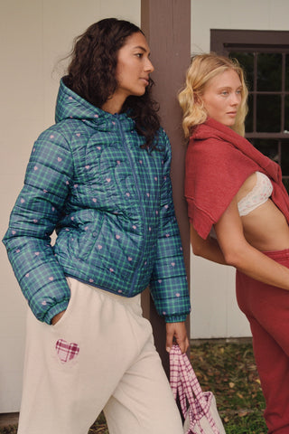 A woman wearing a SOPHISTICA Plaid Hearts Reversible Puffer Jacket, a hooded, zip-front nylon puffer in an emerald green and blue plaid pattern accented by small pink hearts.