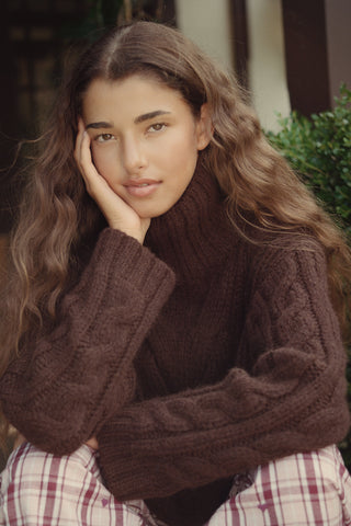 A person wearing a cozy, chocolate brown Naomi Sweater by REVOLUTION KNITS, featuring a cable-knit design and a high turtleneck.