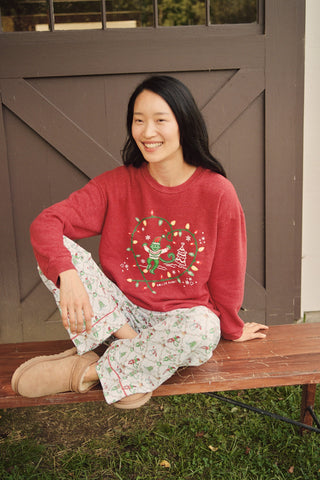 A smiling woman sits on a wooden bench wearing a red Deck The Halls Andie Sweatshirt featuring an illustrated monkey in string lights and patterned Bea Sweatpants.