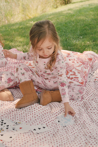 A child in a patterned Kids Seasons Soiree Juniper Pajamas two-piece set and rubber boots sits on a wooden sled and looks through binoculars.