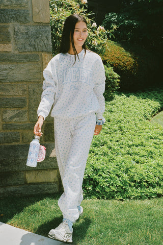 A person wearing a white Roller Rabbit Mini Monkey Blossom Andie Sweatshirt, featuring small blue and green Mini Monkey Blossom prints and large SLEEP Club lettering, stands outdoors, partially in sunlight. Their face is mostly out of frame.