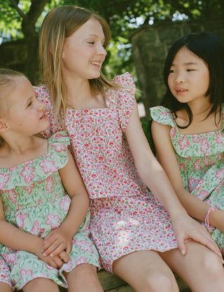 Three young girls sit close together outdoors, each wearing the Roller Rabbit Kids Garden Gate Floral Emmett Top—an ideal choice for girls' summer clothing. They are smiling and appear to be engaged in conversation, with greenery and stone walls in the background.