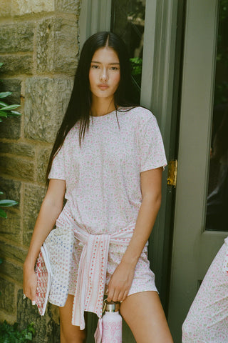 A woman wearing the Roller Rabbit Garden Gate Floral Elena Set stands by a stone wall and glass door, holding a patterned clutch and water bottle, with a matching sweatshirt tied at her waist.
