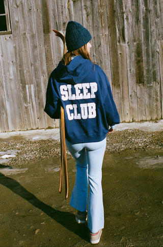 Person wearing a blue jacket with 'Sleep Club' text, standing in front of a wooden wall.