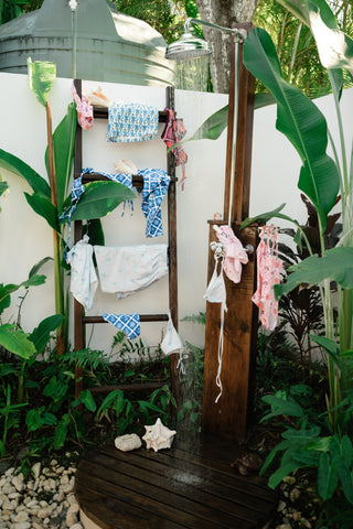 Outdoor shower area with towels and clothes hanging, surrounded by greenery.