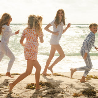 Four children in patterned pajamas run and play barefoot on a sandy beach by the ocean, with gentle waves in the background and sunlight shining through the clouds.