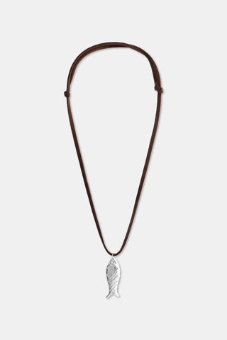 A woman is shown from the shoulders up in a blue and green patterned top, wearing the Roller Rabbit Sardine Cord Necklace featuring a silver fish-shaped pendant on an adjustable brown cord.