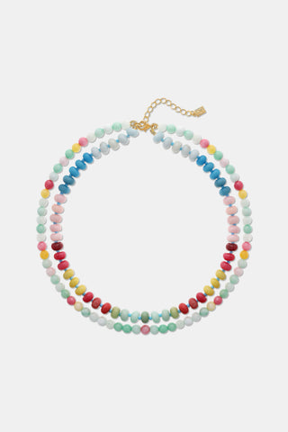 A woman wears Roller Rabbit’s Beaded Necklace Stack—one necklace with pastel beads and a white flower charm, the other with colorful beaded chains—paired with a white knit top featuring floral cutouts. Only her lower face and neck are visible.