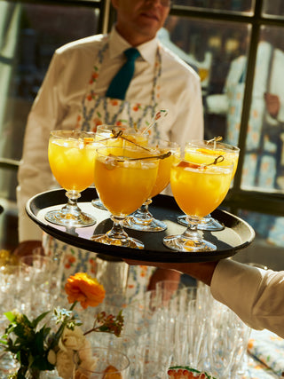 A server holds a tray of orange cocktails with garnishes at a sunlit event, while a guest in a white shirt and blue tie stands in the background, adding a touch of elegance to the scene.