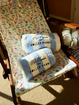 A deck chair with a colorful sailboat print holds two blue and white towels labeled “Kit Kemper for Roller Rabbit.” White cups are stacked nearby on the right, while sunlight brightens the cheerful scene.
