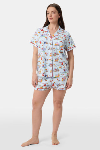 A woman with curly hair smiles in Roller Rabbit Spain Monkey Polo Pajamas—light blue shortie PJs made from 100 Pima cotton with colorful whimsical prints and red piping—standing against a plain white background.