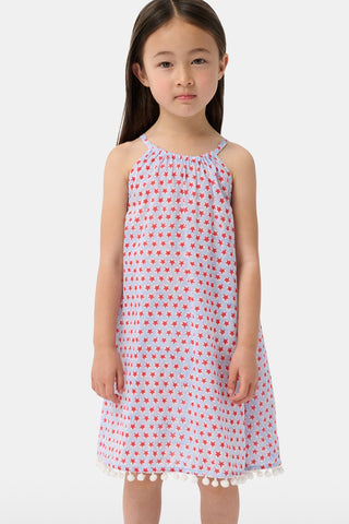 A young girl with long brown hair stands facing forward in the Roller Rabbit Kids Starry Dot Carrie Cover Up—a sleeveless, knee-length white dress featuring a red dotted pattern and pom-pom trim. She has a neutral expression and a light background behind her.