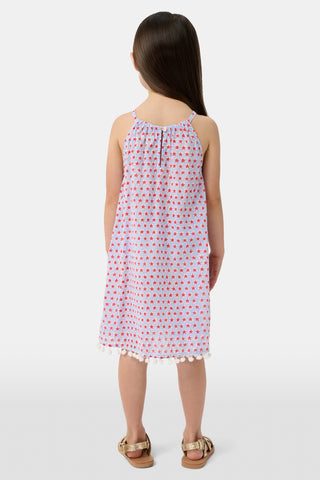 A young girl with long brown hair stands facing forward in the Roller Rabbit Kids Starry Dot Carrie Cover Up—a sleeveless, knee-length white dress featuring a red dotted pattern and pom-pom trim. She has a neutral expression and a light background behind her.