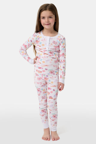 A young girl smiles barefoot in Roller Rabbit Kids Summer Memories Pajamas, made from soft Pima Cotton with playful, colorful prints on white. She stands against a light gray background. Made in Peru.