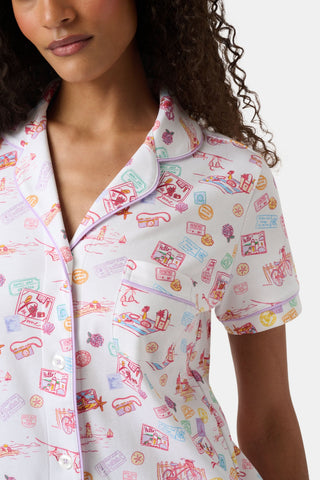 A woman with curly hair models the Roller Rabbit Summer Memories Fiora Pajamas, a Pima cotton set with a colorful sushi print, including a short-sleeve button-up top and long pants, against a white background.
