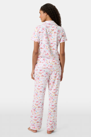 A woman with curly hair models the Roller Rabbit Summer Memories Fiora Pajamas, a Pima cotton set with a colorful sushi print, including a short-sleeve button-up top and long pants, against a white background.