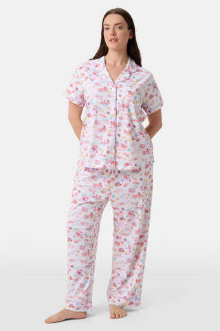 A woman with curly hair models the Roller Rabbit Summer Memories Fiora Pajamas, a Pima cotton set with a colorful sushi print, including a short-sleeve button-up top and long pants, against a white background.