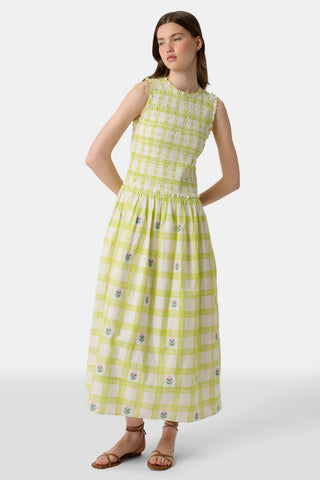 A woman wears the Roller Rabbit Sun Check Blossom Embroidery Emma Dress, a sleeveless green maxi with floral embroidery and smocked bodice, paired with brown sandals. She stands relaxed, looking to the side.