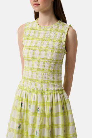 A woman wears the Roller Rabbit Sun Check Blossom Embroidery Emma Dress, a sleeveless green maxi with floral embroidery and smocked bodice, paired with brown sandals. She stands relaxed, looking to the side.