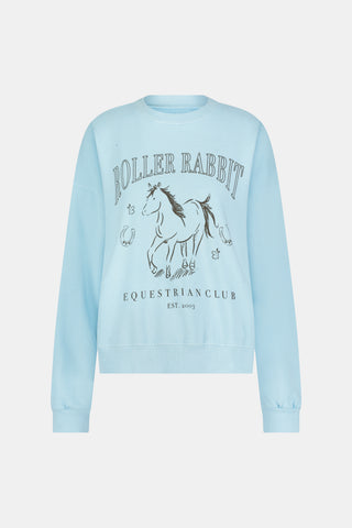 Wearing the Roller Rabbit Equestrian Club Phoebe Sweatshirt in baby blue, a woman pairs it with horse-patterned shorts as she holds a baby goat. She stands by a wooden fence with greenery in the background, smiling softly.