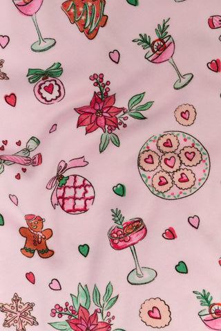 Two women sit on a patterned blanket outdoors wearing matching light pink Seasons Soiree Hayden Pajamas by NETTALCO, featuring a classic Pima cotton top and ruffled shorts decorated with holiday-themed illustrations.