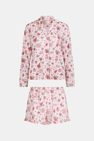 Two women sit on a patterned blanket outdoors wearing matching light pink Seasons Soiree Hayden Pajamas by NETTALCO, featuring a classic Pima cotton top and ruffled shorts decorated with holiday-themed illustrations.