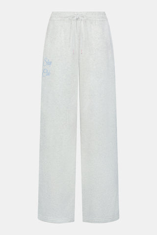 A person is wearing the Roller Rabbit Sleep Club Mimi Sweatpants in light gray, featuring wide legs and crafted from velvety soft Dream Fleece with “Sleep Club” printed in light blue on the left thigh, paired with white sneakers and a cropped white top. The background is plain white.
