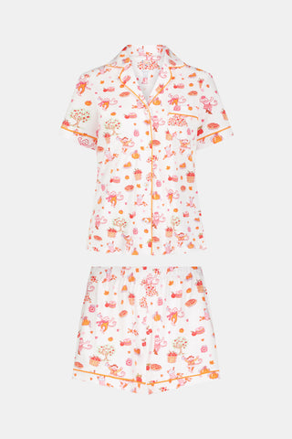 A woman smiles while wearing the Roller Rabbit Sweater Weather Polo Pajamas, a matching set of printed Pima PJs featuring colorful pink and orange cupcakes, flowers, and playful designs on a white background.
