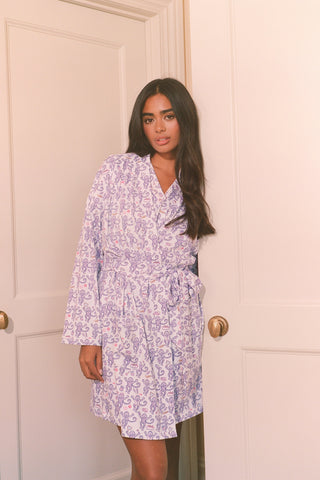 A woman smiles while wearing the TEXPIMA Get Ready With Me Blake Robe, featuring a knee-length cut and a purple and pink paisley pattern, tied at the waist, against a plain white background.