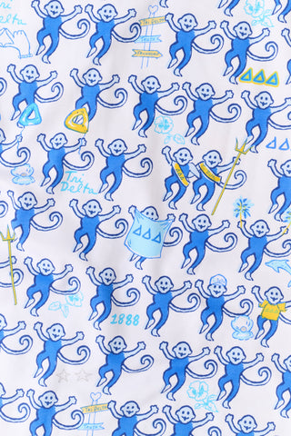 A woman wearing blue and white patterned Tri Delta Monkey Polo Pajamas by Roller Rabbit holds a large striped bag. She stands outdoors near a brick wall and green plants, with sunlight filtering through the leaves.
