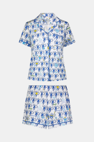 A woman wearing blue and white patterned Tri Delta Monkey Polo Pajamas by Roller Rabbit holds a large striped bag. She stands outdoors near a brick wall and green plants, with sunlight filtering through the leaves.