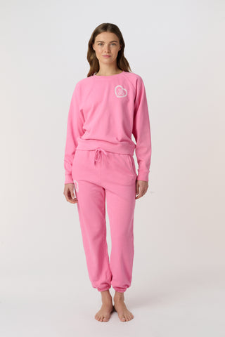 A woman wears the Roller Rabbit Vintage Valentine Crewneck, a pink pullover sweatshirt with white heart-shaped designs, standing against a plain light background and smiling softly.