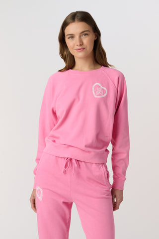 A woman wears the Roller Rabbit Vintage Valentine Crewneck, a pink pullover sweatshirt with white heart-shaped designs, standing against a plain light background and smiling softly.