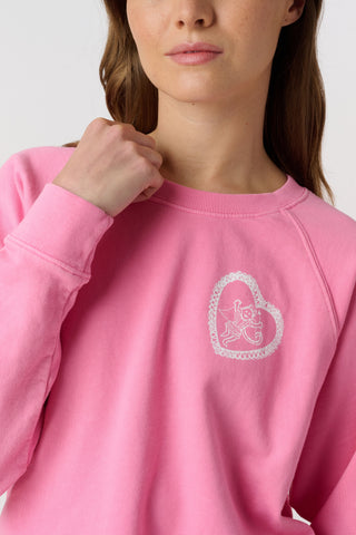 A woman wears the Roller Rabbit Vintage Valentine Crewneck, a pink pullover sweatshirt with white heart-shaped designs, standing against a plain light background and smiling softly.