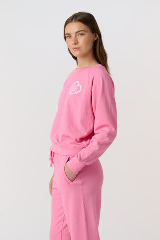 A woman wears the Roller Rabbit Vintage Valentine Crewneck, a pink pullover sweatshirt with white heart-shaped designs, standing against a plain light background and smiling softly.