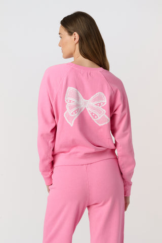 A woman wears the Roller Rabbit Vintage Valentine Crewneck, a pink pullover sweatshirt with white heart-shaped designs, standing against a plain light background and smiling softly.