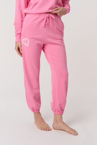 A person wears the Roller Rabbit Vintage Valentine Sweatpants—pink fleece with a drawstring waist and white heart designs—standing barefoot against a plain light background, showcasing only the lower half of their body.