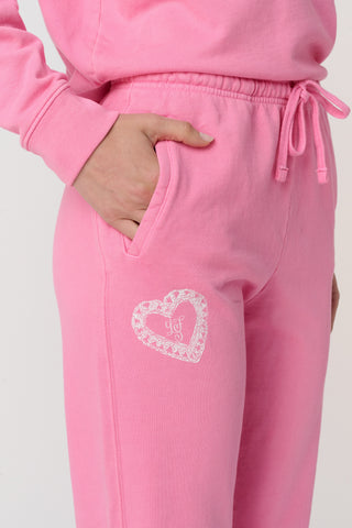 A person wears the Roller Rabbit Vintage Valentine Sweatpants—pink fleece with a drawstring waist and white heart designs—standing barefoot against a plain light background, showcasing only the lower half of their body.